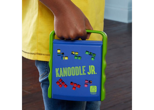 A person holding a blue and green Kanooodle Jr. toy in their hand, wearing a yellow shirt and blue jeans, with a dark wood floor in the background.